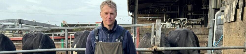 a man wearing overalls stood in a farmyard with cows in the background